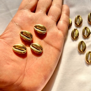 May include: A collection of gold cowrie shells, some are in a hand, others are scattered on a white surface.
