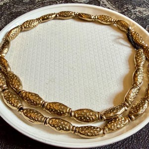 May include: A gold-coloured necklace featuring oval beads. The beads are strung on a black cord and presented on a white plate with a textured surface. The necklace has a vintage or antique appearance.