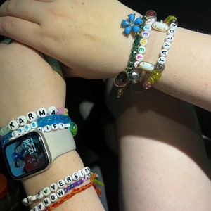 May include: A person's wrist with multiple beaded bracelets. The bracelets spell out "KARMA", "IF", "TAYLOR", "LOVE", and "AKNOW". The person is also wearing a smartwatch with a photo of three people on the screen.