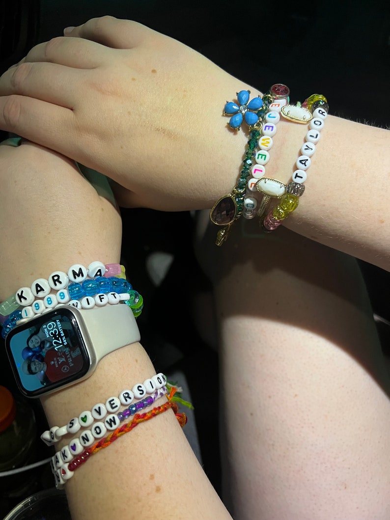May include: A person's wrist with multiple beaded bracelets. The bracelets spell out "KARMA", "IF", "TAYLOR", "LOVE", and "AKNOW". The person is also wearing a smartwatch with a photo of three people on the screen.