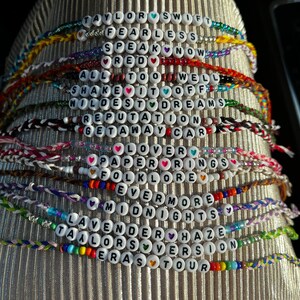 May include: A stack of colorful bracelets with white beads spelling out the names of Taylor Swift's albums, including "Taylor Swift", "Fearless", "Speak Now", "Red", "1989", "Reputation", "Lover", "Folklore", "Evermore", "Midnights", "Taylor's Version", and "Eras Tour".