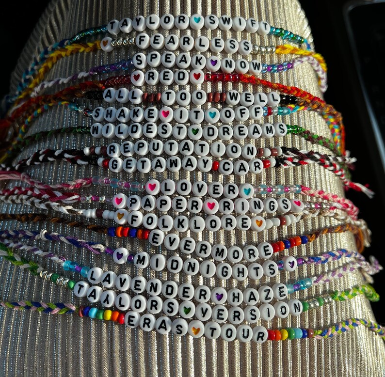 May include: A stack of colorful bracelets with white beads spelling out the names of Taylor Swift's albums, including "Taylor Swift", "Fearless", "Speak Now", "Red", "1989", "Reputation", "Lover", "Folklore", "Evermore", "Midnights", "Taylor's Version", and "Eras Tour".