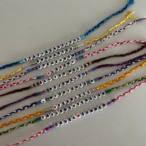May include: A collection of colorful friendship bracelets with different song titles from Taylor Swift's albums, including "Stay Beautiful", "Enchanted", "Speak Now", "Lover", "All Too Well", "Bejeweled", "Midnights", "Fearless", "Taylor's Version", and "Cruel Summer".
