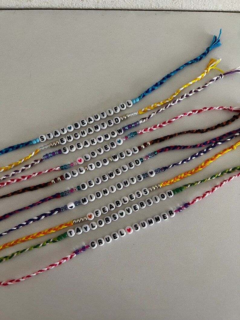 May include: A collection of colorful friendship bracelets with different song titles from Taylor Swift's albums, including "Stay Beautiful", "Enchanted", "Speak Now", "Lover", "All Too Well", "Bejeweled", "Midnights", "Fearless", "Taylor's Version", and "Cruel Summer".