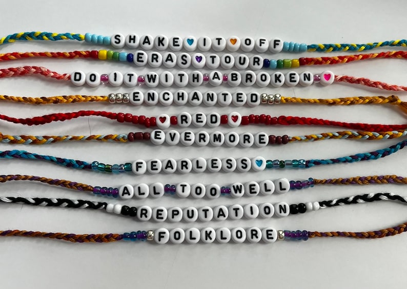 May include: Ten colorful friendship bracelets with different Taylor Swift album titles spelled out in white beads. The bracelets are made with colorful embroidery floss.