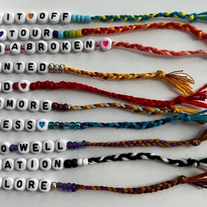 May include: Ten colorful friendship bracelets with white beads spelling out the names of Taylor Swift albums: "Shake It Off", "1989", "Reputation", "Red", "Fearless", "Speak Now", "Lover", "Folklore", "Evermore", and "All Too Well".