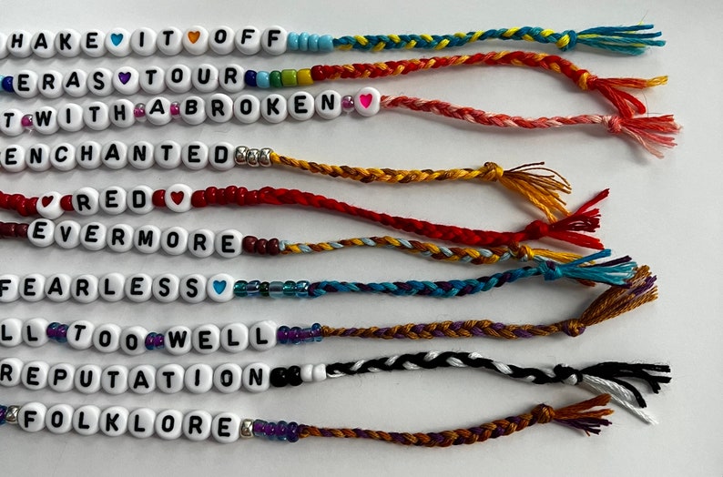 May include: Ten colorful friendship bracelets with white beads spelling out the names of Taylor Swift albums: "Shake It Off", "1989", "Reputation", "Red", "Fearless", "Speak Now", "Lover", "Folklore", "Evermore", and "All Too Well".