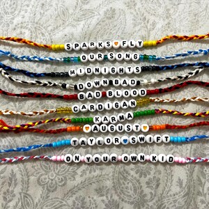 May include: A set of 10 friendship bracelets with colorful string and white beads spelling out song titles by Taylor Swift. The titles are "Sparks Fly", "Our Song", "Midnights", "Down Bad", "Bad Blood", "Cardigan", "Karma", "August", "Taylor Swift", and "On Your Own Kid".
