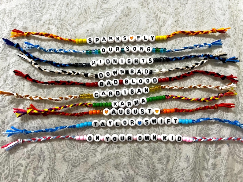 May include: A set of 10 friendship bracelets with colorful string and white beads spelling out song titles by Taylor Swift. The titles are "Sparks Fly", "Our Song", "Midnights", "Down Bad", "Bad Blood", "Cardigan", "Karma", "August", "Taylor Swift", and "On Your Own Kid".
