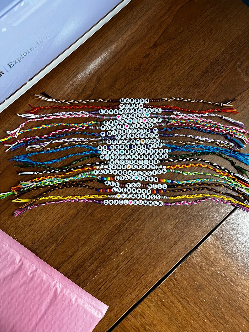 May include: A collection of 20 colorful friendship bracelets with beaded words, including "Maroon", "Red", "Lavender Haze", "Lover", "Shake It Off", "Paper Rings", "Midnights", "Speak Now", "Get Away Car", "Stay Beautiful", "On Your Own Kid", "Wildest Dreams", "Reputation", "Love Story", "Taylor Swift", "Eras Tour", "All Too Well", "Evermore", "Fearless", "Cruel Summer".