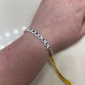 May include: A yellow and gold beaded bracelet with the word "Fearless" spelled out in black letters on white beads.