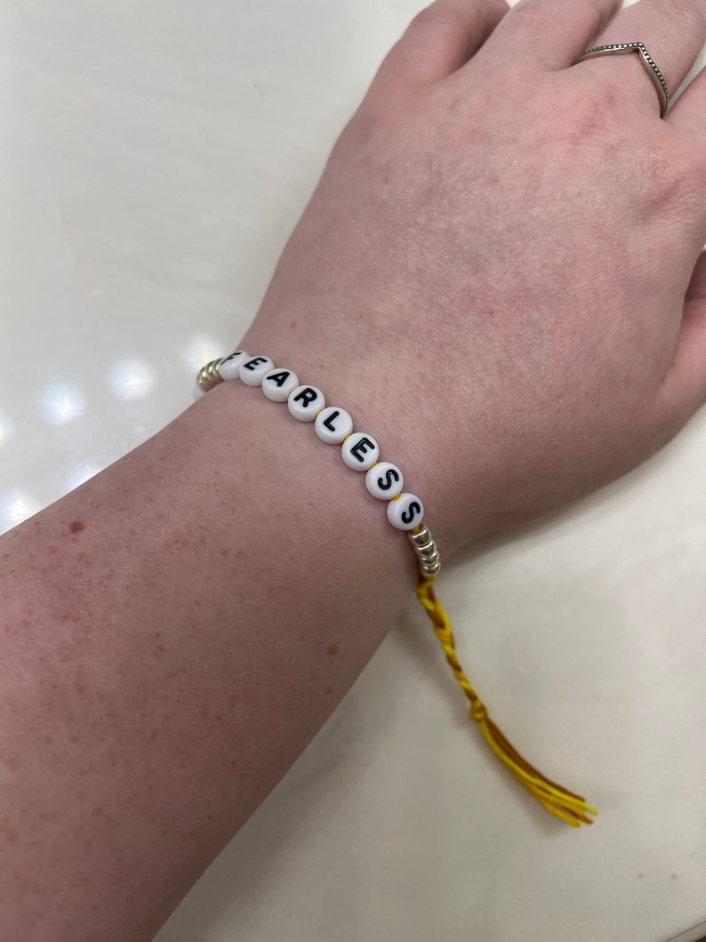 May include: A yellow and gold beaded bracelet with the word "Fearless" spelled out in black letters on white beads.