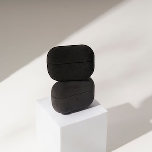 Puede incluir: Dos estuches negros de tela suave apilados uno encima del otro. Los estuches están sobre un pedestal cuadrado blanco.