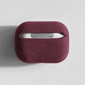 Puede incluir: Una funda de color burdeos suave y peluda para auriculares inal&aacute;mbricos. La funda tiene una forma rectangular y una parte superior e inferior ligeramente redondeadas.