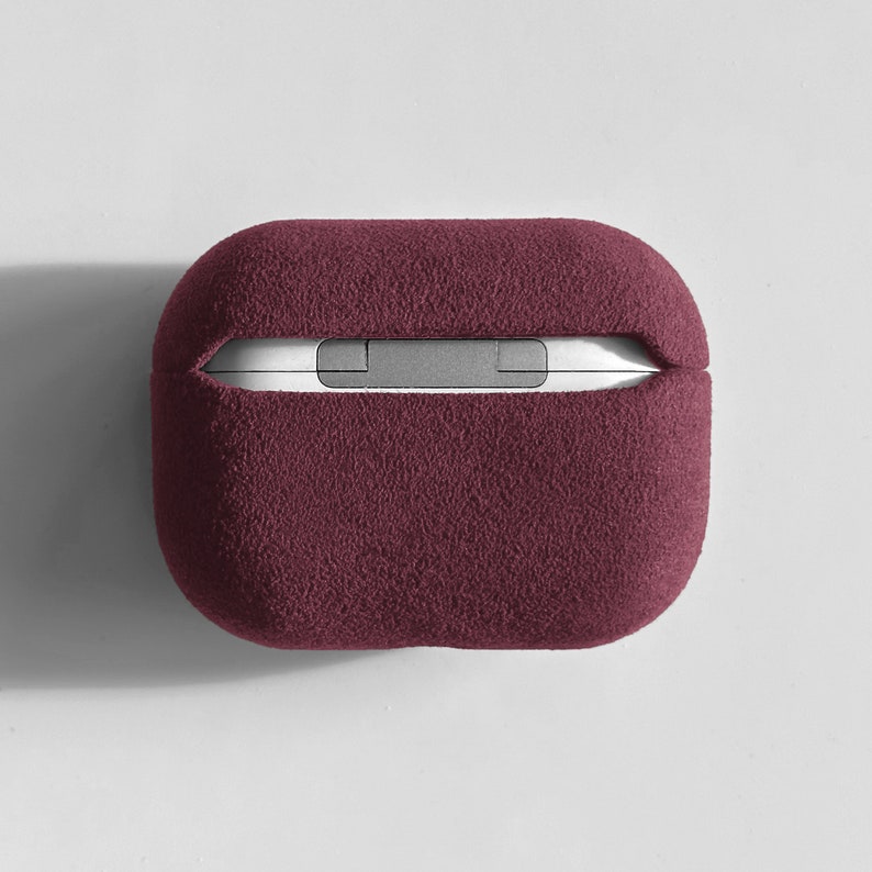 Puede incluir: Una funda de color burdeos suave y peluda para auriculares inal&aacute;mbricos. La funda tiene una forma rectangular y una parte superior e inferior ligeramente redondeadas.