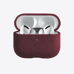 Puede incluir: Una funda protectora de color rojo burdeos para Apple AirPods Pro con un acabado de tejido texturizado. La funda est&aacute; abierta, revelando los AirPods blancos en el interior.