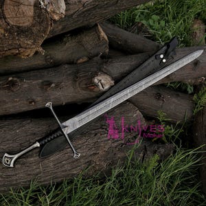 May include: A silver sword with a black leather handle and scabbard. The sword is lying on a pile of logs with green grass in the background.