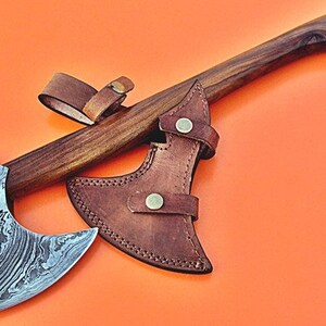 May include: A hand-forged Damascus steel axe with a wooden handle and a brown leather sheath. The axe has a unique, swirling pattern on the blade.