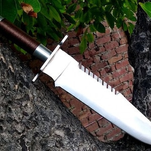 May include: A large, silver knife with a brown leather handle. The knife has a serrated edge and a pointed tip. The knife is lying on a tree trunk.