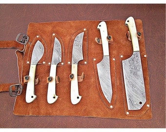 May include: A set of five knives with white handles and patterned blades are displayed in a brown leather roll. The roll has five slots for the knives and is secured with a buckle.