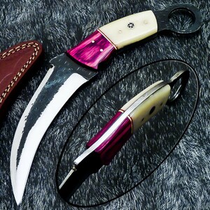 May include: A curved karambit knife with a polished silver blade and a handle featuring cream, magenta, and black accents. The handle has a ring at the end. A leather sheath is visible in the background.
