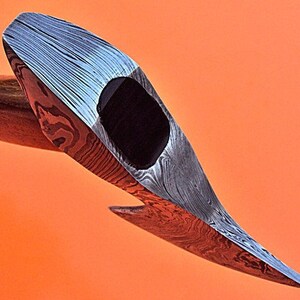 May include: A close-up of a hand-forged axe head with a Damascus steel blade. The blade has a distinctive pattern of swirling lines and a black epoxy handle.