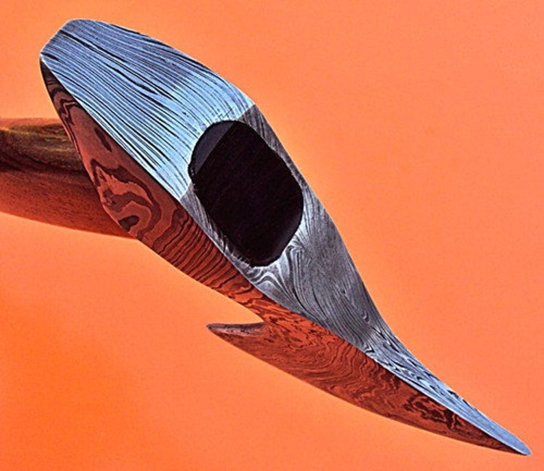 May include: A close-up of a hand-forged axe head with a Damascus steel blade. The blade has a distinctive pattern of swirling lines and a black epoxy handle.