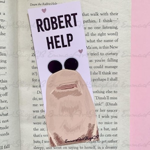 以下が含まれることがあります： 「ROBERT HELP」の文字と漫画のキャラクターが描かれた、気まぐれなブックマーク。ブックマークは開いた本の中に置かれ、テキストと画像の遊び心のあるコントラストを生み出しています。