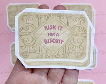 Può includere: Un adesivo a forma di biscotto con la scritta "RISK IT FOR A BISCUIT". L'adesivo è marrone chiaro con un bordo bianco e decorazioni a volute. L'adesivo è tenuto in mano su sfondo rosa.