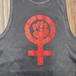 K&ouml;nnte beinhalten: Schwarzes Tanktop mit einem roten Glitzer-Feministensymbol mit einer Faust in einem Kreis.