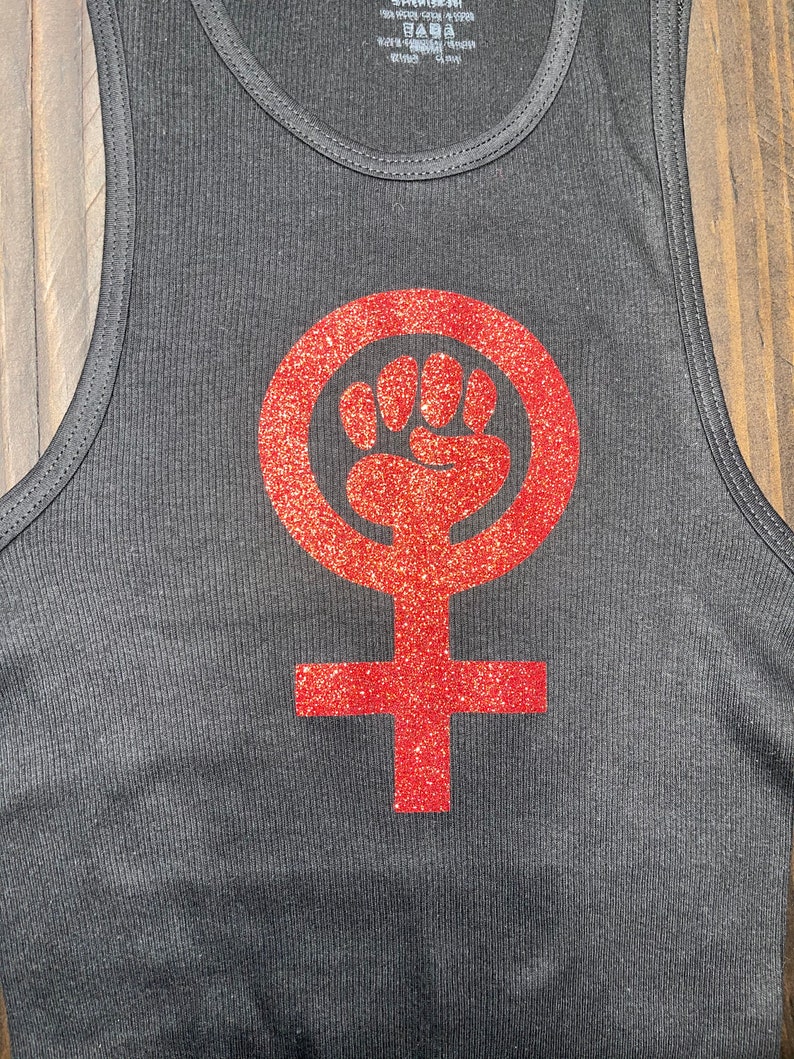 K&ouml;nnte beinhalten: Schwarzes Tanktop mit einem roten Glitzer-Feministensymbol mit einer Faust in einem Kreis.