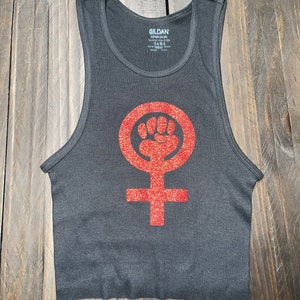 K&ouml;nnte beinhalten: Ein schwarzes Tanktop mit einem roten Glitzer-Feministensymbol. Das Symbol ist ein Kreis mit einer erhobenen Faust darin und einem Kreuz darunter.