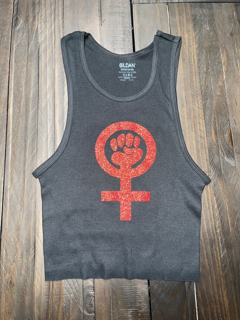K&ouml;nnte beinhalten: Ein schwarzes Tanktop mit einem roten Glitzer-Feministensymbol. Das Symbol ist ein Kreis mit einer erhobenen Faust darin und einem Kreuz darunter.