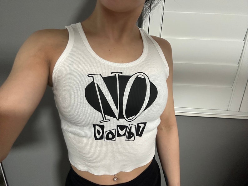 Op de afbeelding: Wit getailleerd tanktop met zwarte tekst die "NO DOUBT" luidt in een gestileerd lettertype.