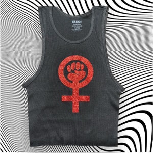 Puede incluir: Un tanque gris oscuro con un símbolo feminista rojo de purpurina que presenta un puño levantado dentro de un círculo.