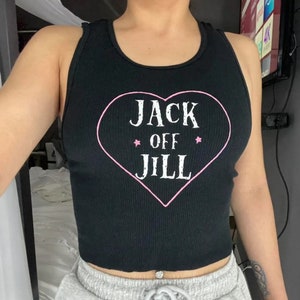 K&ouml;nnte beinhalten: Schwarzes geripptes Tanktop mit einem rosa Herz-Grafik, das den wei&szlig;en Text "JACK OFF JILL" tr&auml;gt. Das Tanktop hat einen Rundhalsausschnitt und ein Racerback-Design. Die Grafik enth&auml;lt zwei rosa Sterne.