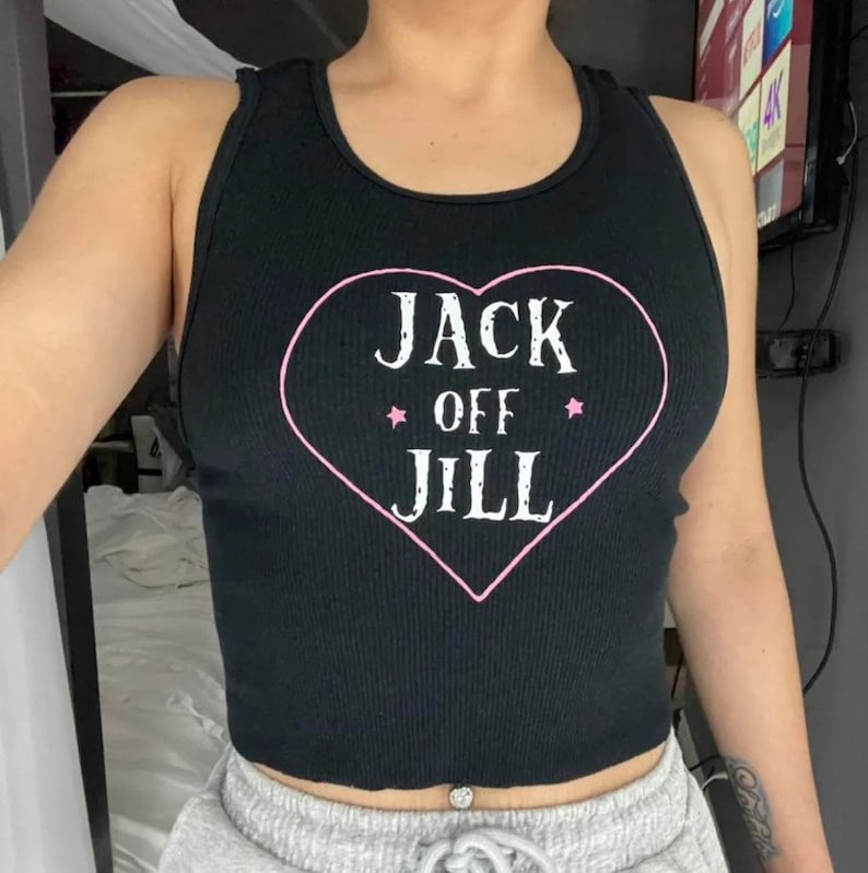 K&ouml;nnte beinhalten: Schwarzes geripptes Tanktop mit einem rosa Herz-Grafik, das den wei&szlig;en Text "JACK OFF JILL" tr&auml;gt. Das Tanktop hat einen Rundhalsausschnitt und ein Racerback-Design. Die Grafik enth&auml;lt zwei rosa Sterne.