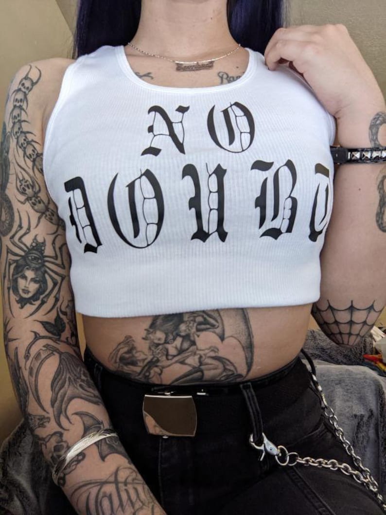 以下が含まれることがあります： 黒いゴシック体で「NO DOUBT」とプリントされた白いタンクトップ。タンクトップはクロップド丈で、リブの質感があります。