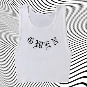 May include: White ribbed tank top with the word "GWEN" in black gothic lettering. The top has a scoop neck and a cropped fit. The background features a black and white wavy pattern.