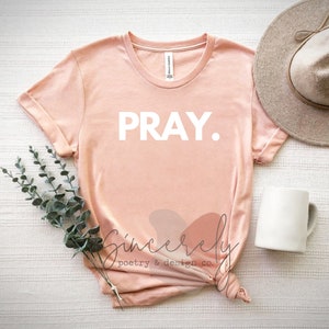 May include: A light peach-colored t-shirt with the word "PRAY." in white block letters. The shirt is styled with a neutral-toned hat, a white mug, and sprigs of greenery. The text "Sincerely poetry & design co." is also visible.