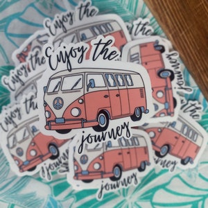 May include: A collection of stickers featuring a vintage-style camper van in shades of coral, white, and blue. The stickers have the text "Enjoy the journey" in a handwritten font. The stickers are scattered on a teal and white patterned background.