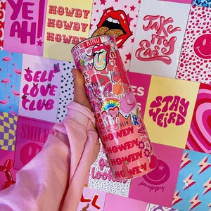 Puede incluir: Un vaso rosa y blanco cubierto de pegatinas, incluyendo un arcoíris y la palabra "HOWDY". El fondo presenta carteles rosas con frases como "YEAH!" y "SELF LOVE CLUB".
