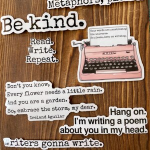 May include: A collection of white stickers with black text on a wooden background. The stickers feature phrases like "Be kind," "Read. Write. Repeat," and "Writers gonna write." A pink typewriter sticker is also present.