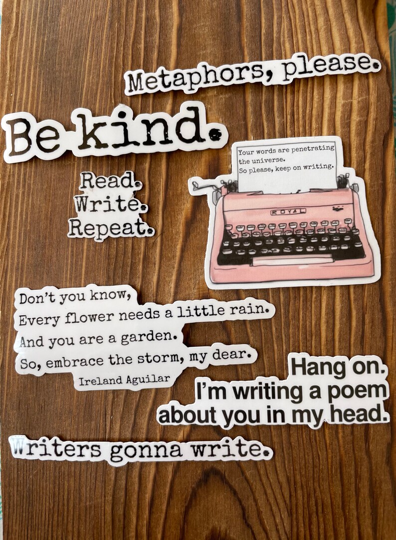 May include: A collection of white stickers with black text on a wooden background. The stickers feature phrases like "Be kind," "Read. Write. Repeat," and "Writers gonna write." A pink typewriter sticker is also present.