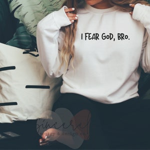 May include: White long-sleeve sweatshirt with the text "I FEAR GOD, BRO." in black. The sweatshirt is made of a soft material. The image also shows a white pillow with black lines and a pillow with a green and white leaf pattern.