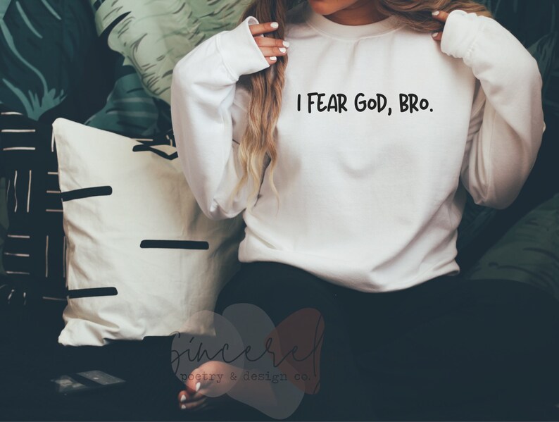 May include: White long-sleeve sweatshirt with the text "I FEAR GOD, BRO." in black. The sweatshirt is made of a soft material. The image also shows a white pillow with black lines and a pillow with a green and white leaf pattern.