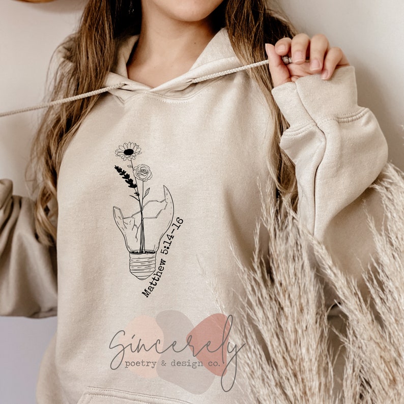 Puede incluir: Sudadera con capucha beige con un dise&ntilde;o floral dentro de una bombilla rota, con el texto "Matthew 5:14-16". La sudadera tambi&eacute;n presenta el texto "Sincerely poetry & design co." en la parte inferior. Tiene una capucha con cord&oacute;n y un bolsillo de canguro.