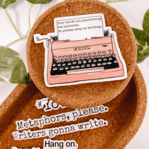 May include: A sticker featuring a pink typewriter with the word "ROYAL" on it, and a piece of paper with the text "Your words are penetrating the universe. So please, keep on writing." The sticker is on a brown cork coaster. Additional stickers with writing are also visible.