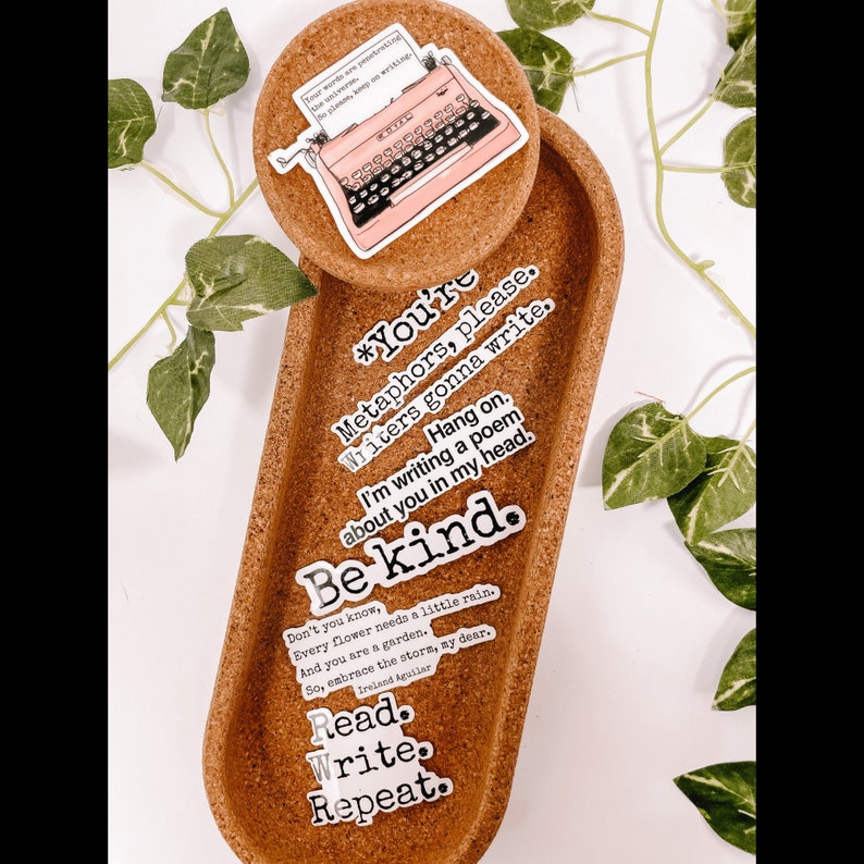 May include: A cork tray and round coaster hold several stickers with writing-themed quotes and a pink typewriter illustration. The stickers include phrases like "Be kind" and "Read. Write. Repeat."