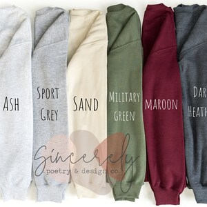 May include: A selection of eight different colored sweatshirts, including white, ash, sport grey, sand, military green, maroon, dark heather, and black. Each sweatshirt has the color name printed on it in a handwritten font. The brand name "Sincerely poetry & design co." is also visible.